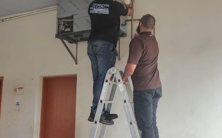 Dois profissionais, um deles sobre escada, trabalhando em equipamento fixado na parede